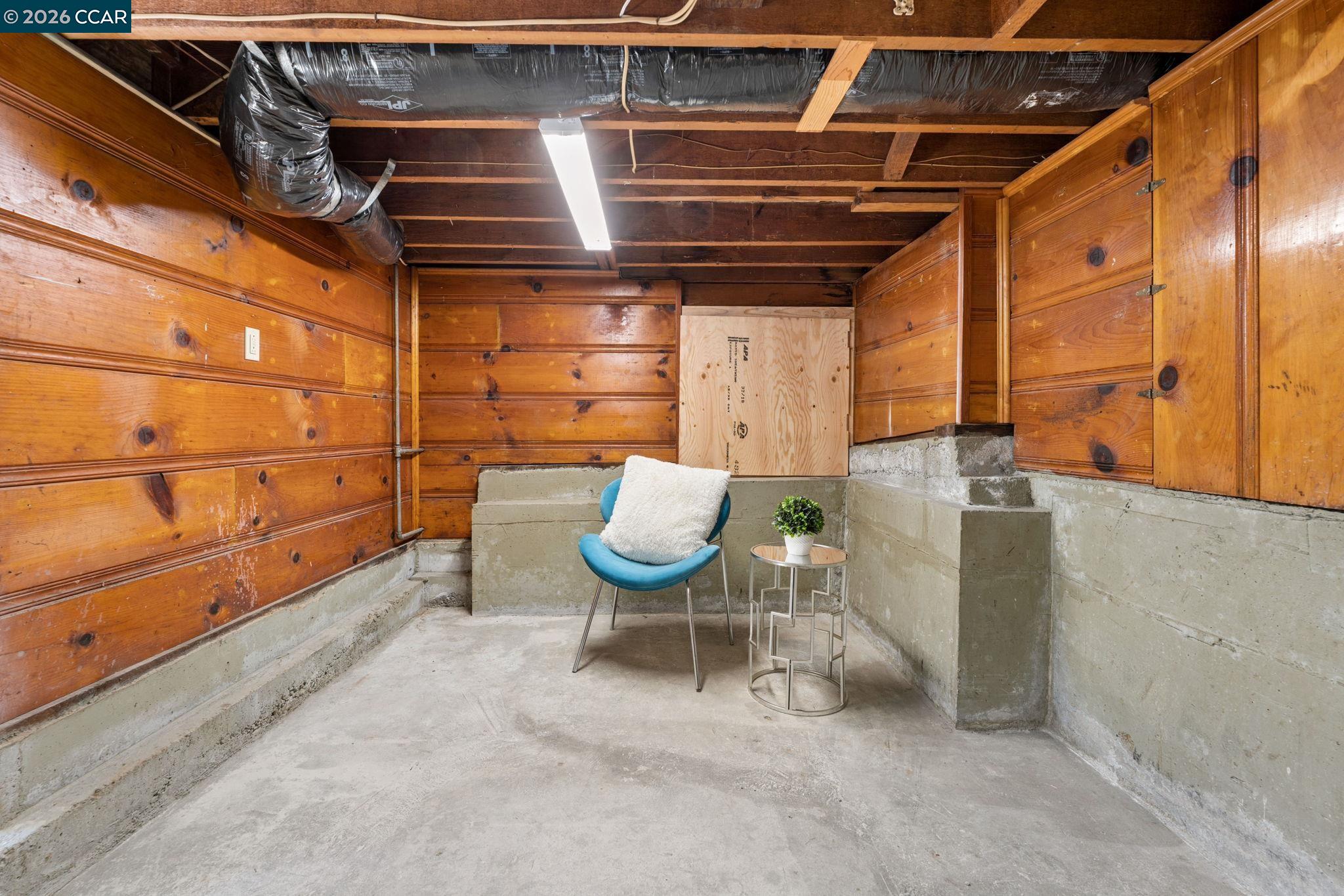 Staged basement with a blue chair, exposed wood walls, concrete floor, and open ceiling.