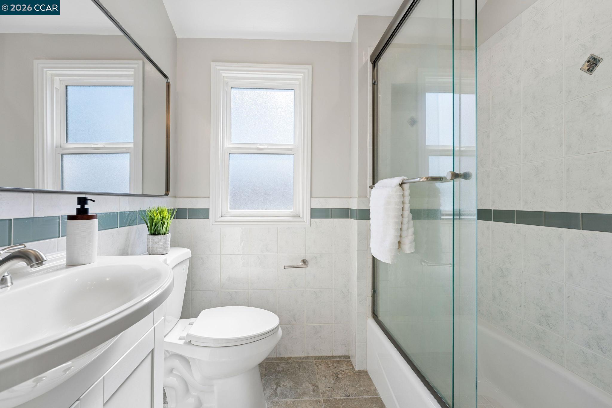Bathroom with a glass-enclosed tub, white vanity, toilet, and blue-green tile accent.