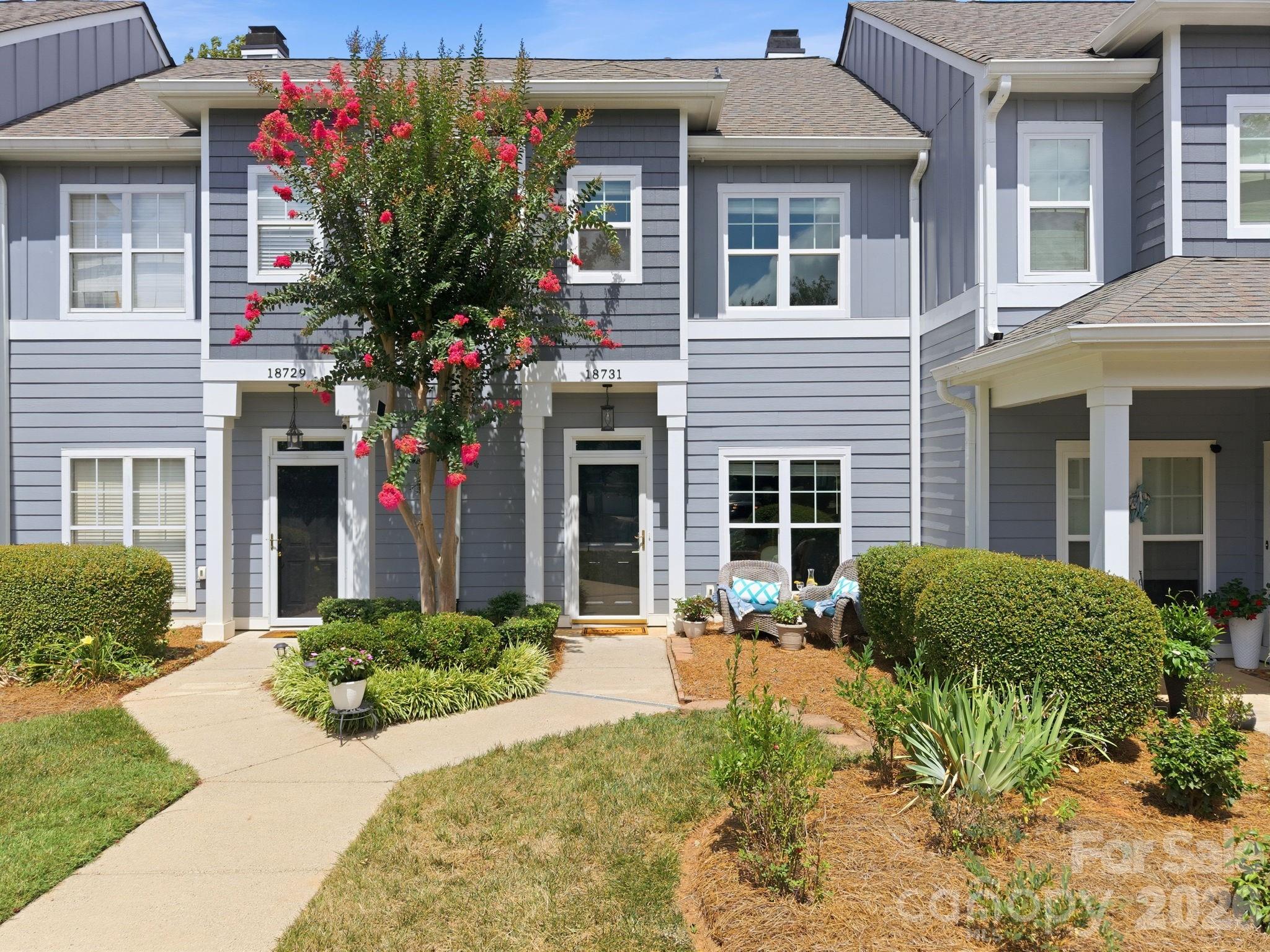 Gray attached home marked 18731 with a flowering tree, landscaped front yard, and patio chairs.