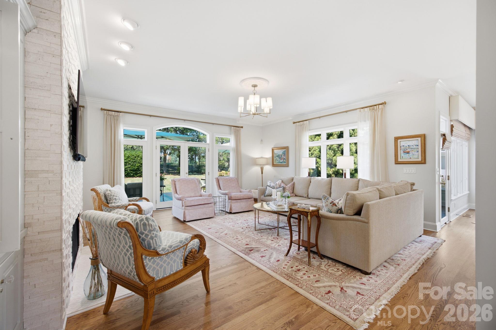 Furnished living room with a stone fireplace, beige sectional, hardwood floors, and tall windows facing the yard.