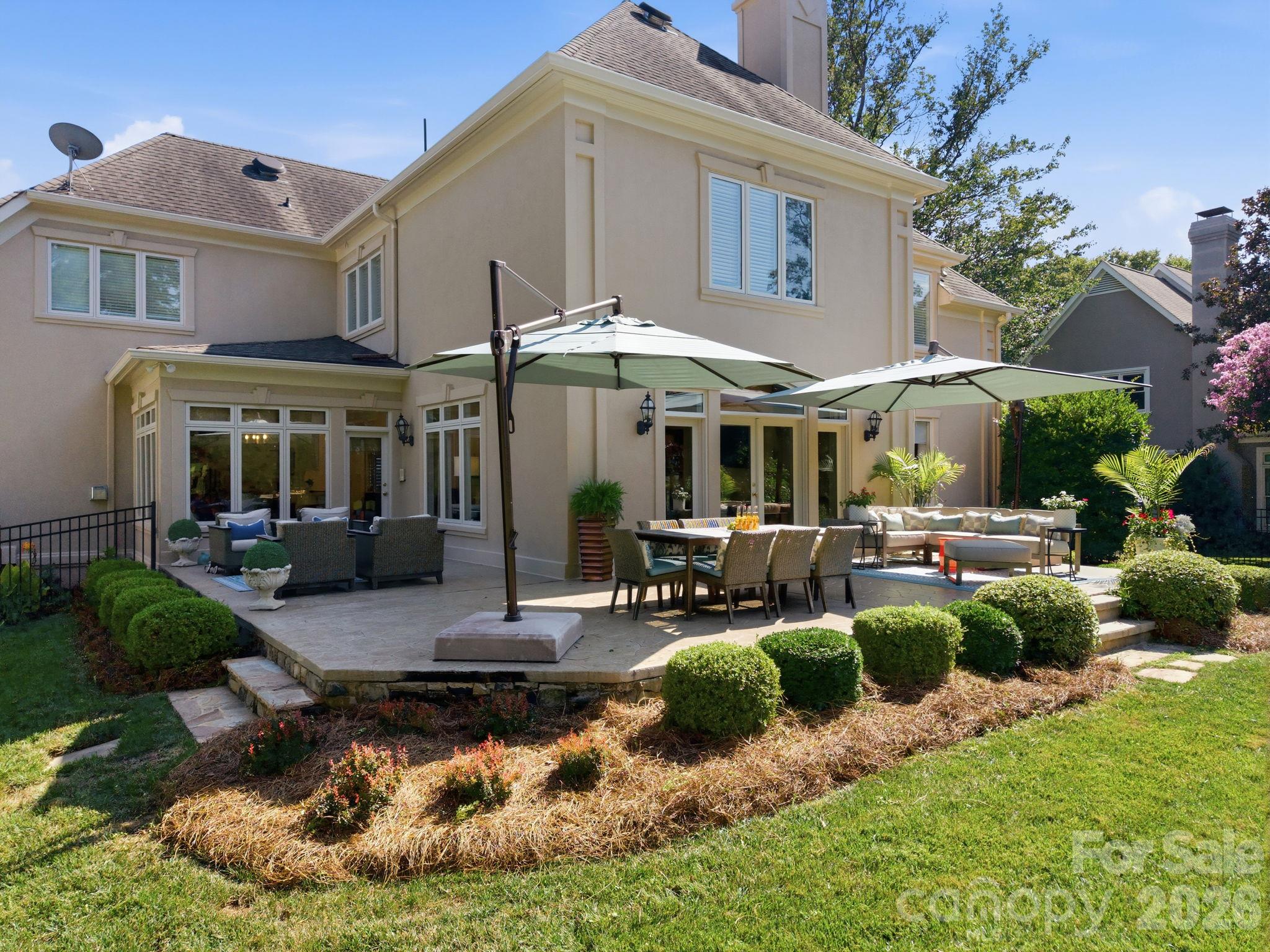Two-story beige home with a furnished stone patio, outdoor dining area, and shade umbrellas.