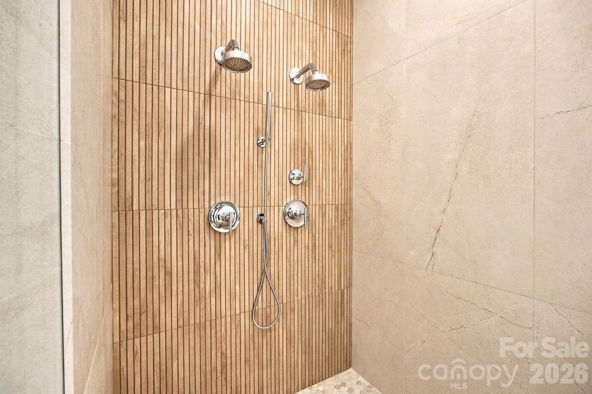 Walk-in shower with dual showerheads, handheld sprayer, and warm vertical wood-look tile.