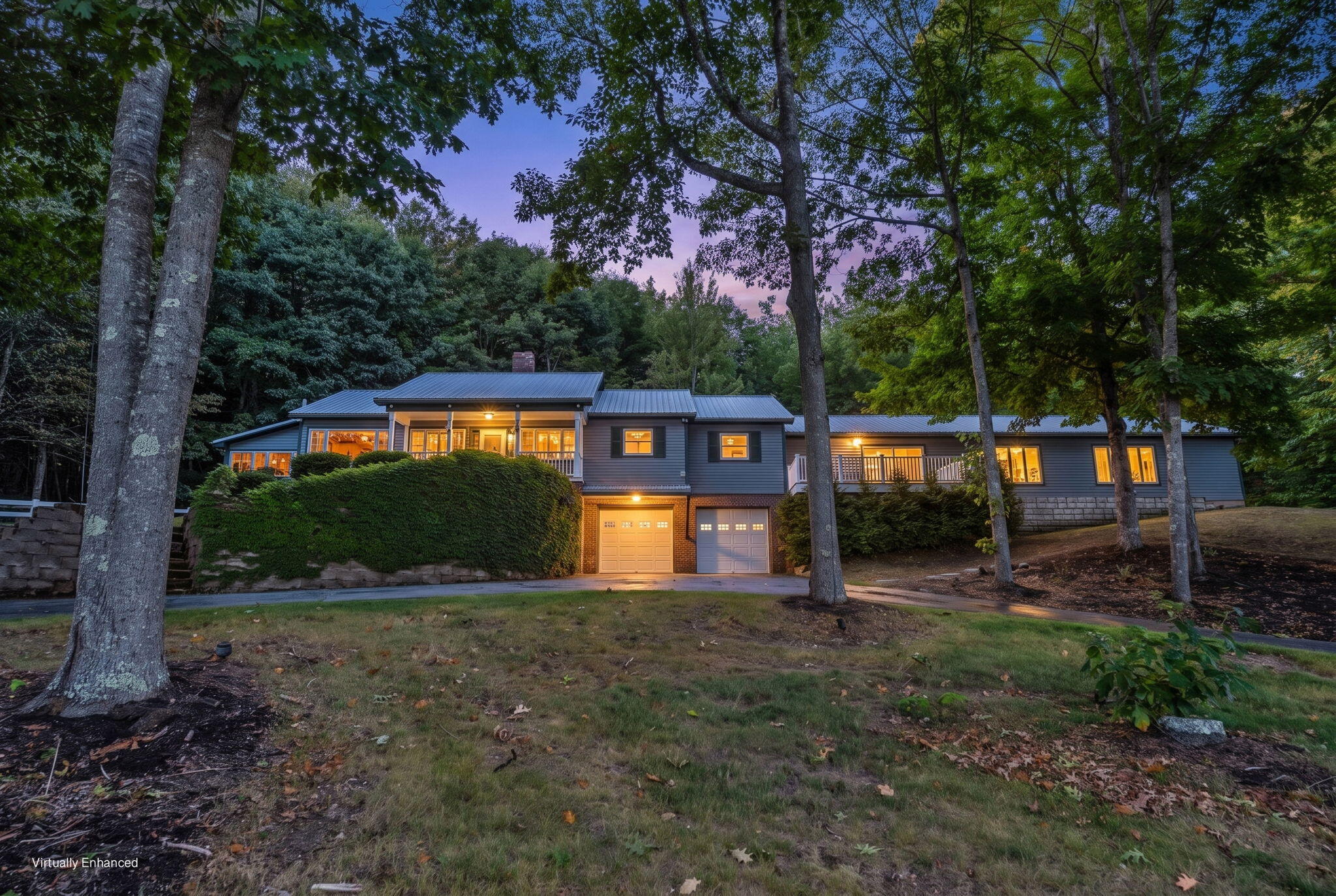 Long, tree-surrounded home at dusk with a covered porch, metal roof, and two illuminated garage doors.
