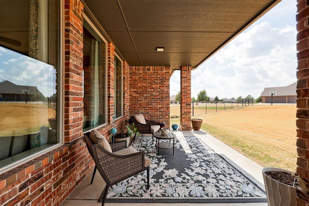 Covered brick patio with wicker seating and a floral rug overlooking a fenced yard.