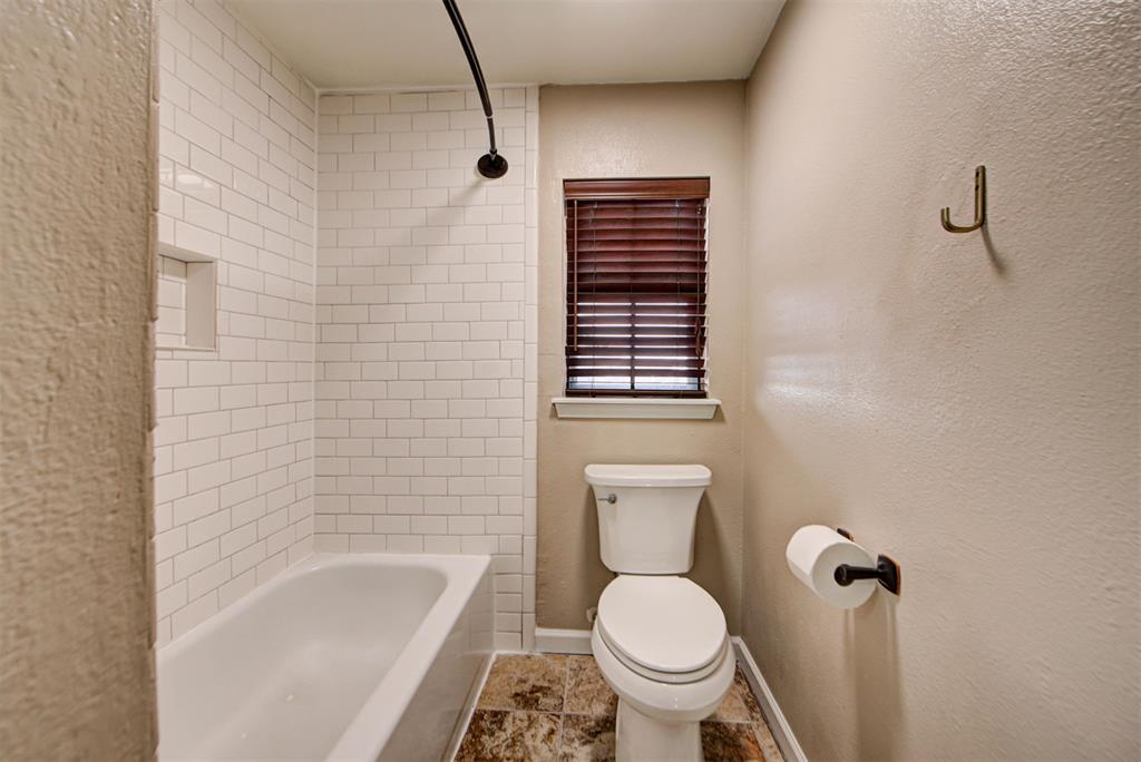 Bathroom with a white subway-tile tub surround, toilet beneath a window, and mottled brown floor tile.