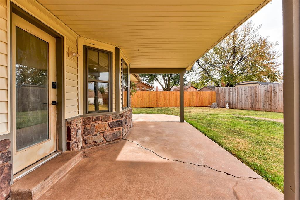 Covered concrete patio beside tan siding and stone veneer, overlooking a fenced grassy backyard.
