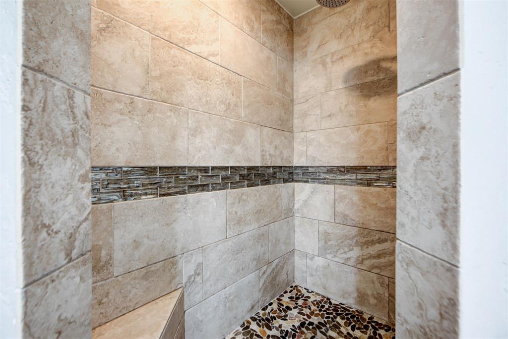 Walk-in shower with beige stone-look tile, a dark mosaic accent band, and pebble flooring.