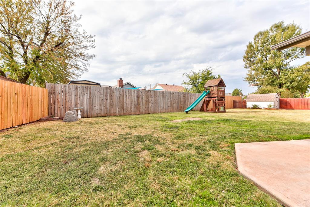 Fenced backyard with a wooden playset, green slide, lawn, and concrete patio.