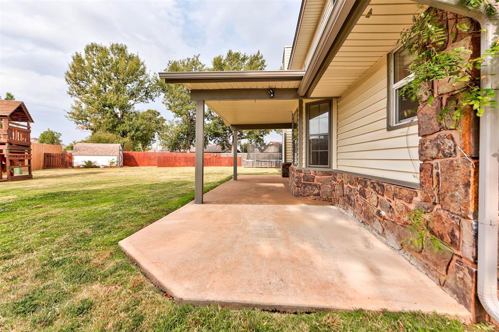 Covered concrete patio beside a stone-accented house, overlooking a broad fenced lawn.