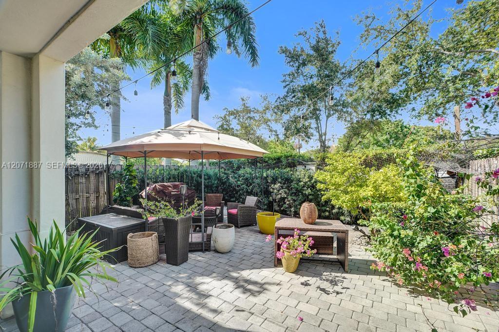 Paved patio with wicker seating beneath a large umbrella, surrounded by palms and flowering plants.
