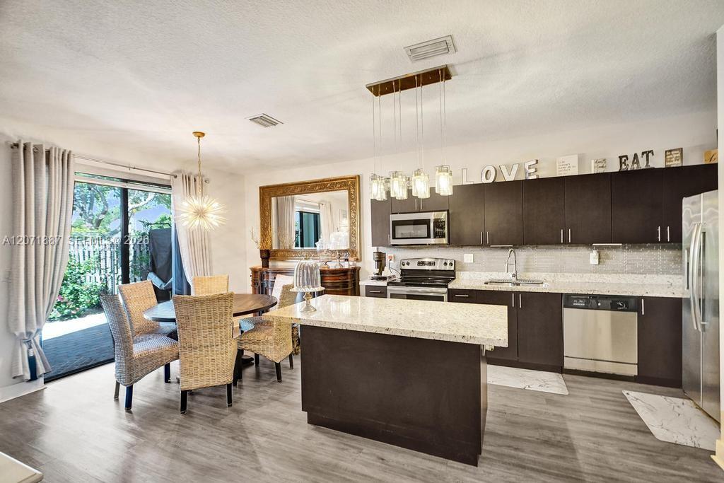 Kitchen with dark cabinetry, stainless steel appliances, a large island, and wicker dining furniture by a sliding glass door.