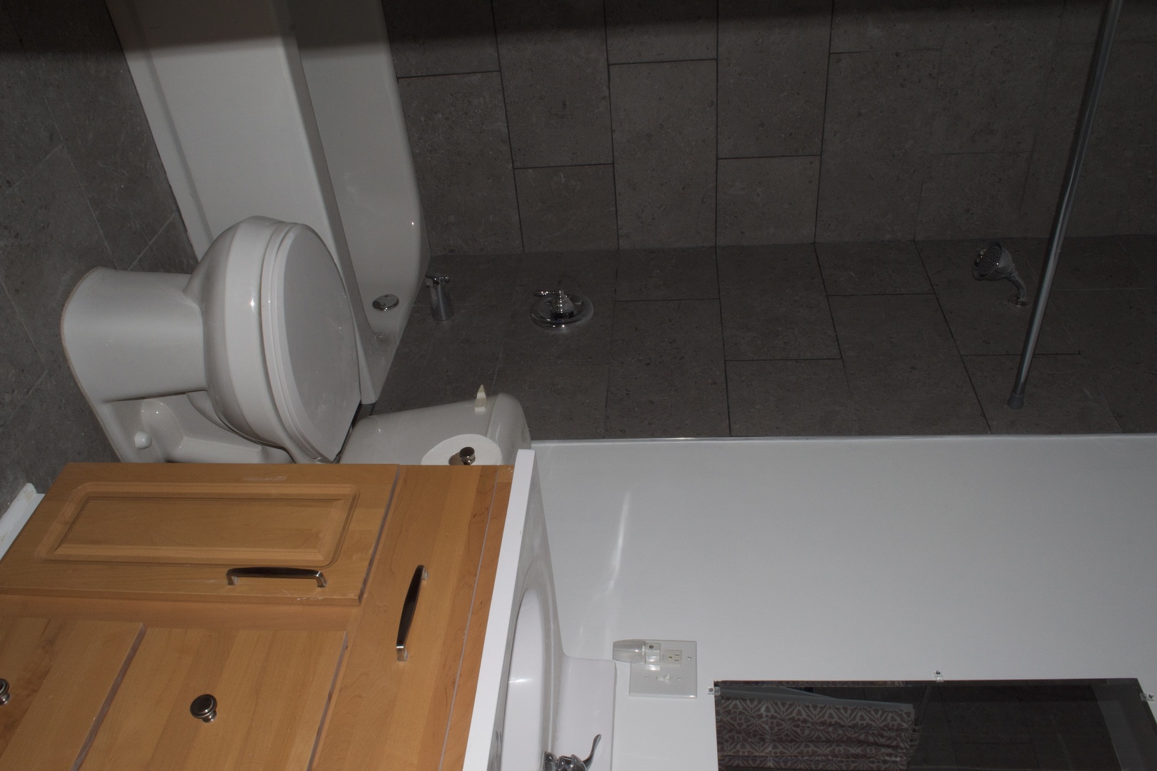 Bathroom with a white tub, toilet, wood vanity, and dark tiled shower.