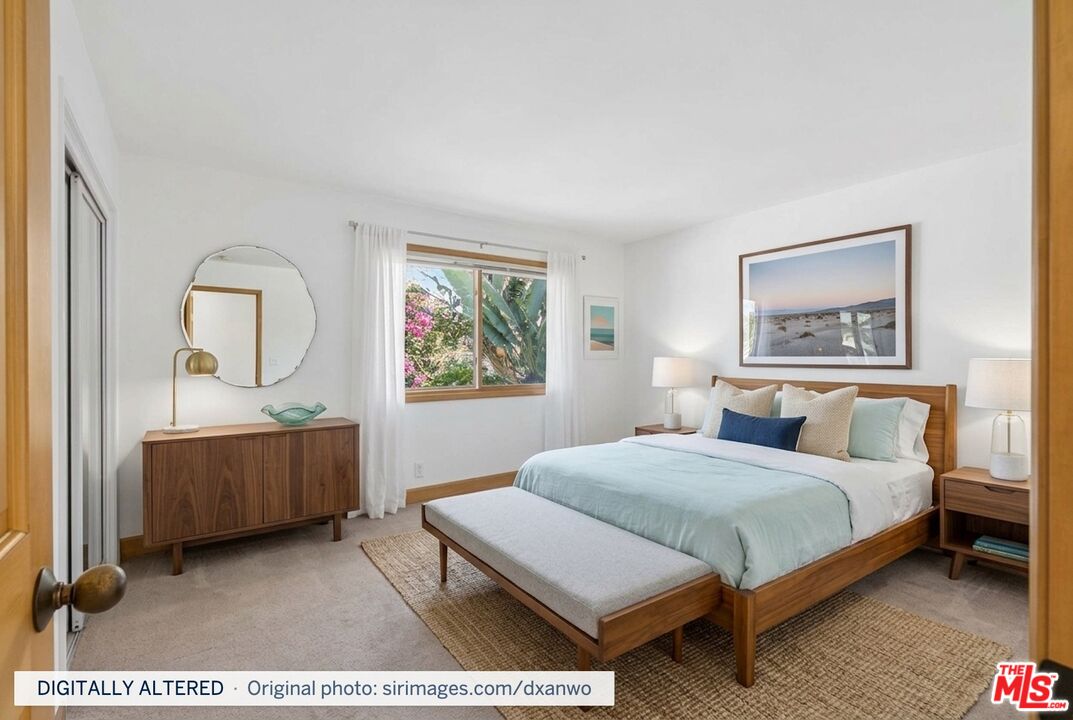 Bedroom with pale blue bedding, wood furniture, and a window overlooking tropical foliage.