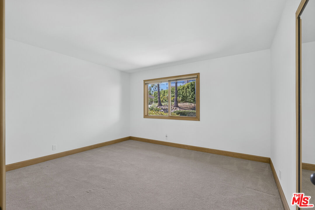Empty carpeted room with wood trim and a window overlooking landscaped greenery.
