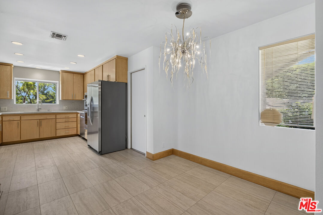 Kitchen with light wood cabinets, stainless steel refrigerator, and a sculptural chandelier over an open tiled area.