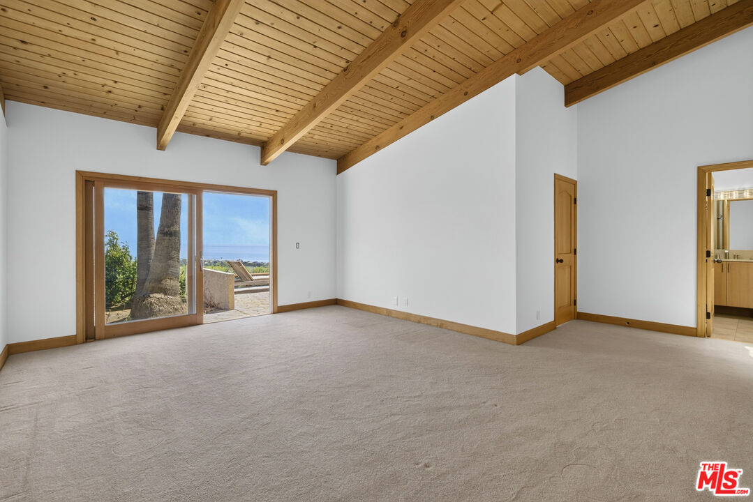 Empty living room with exposed wood beams and sliding glass doors overlooking a patio and ocean.