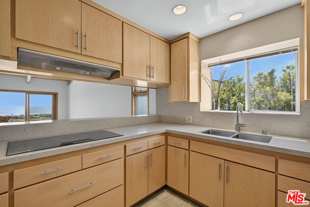 Kitchen with light wood cabinets, pale stone counters, and windows showing trees and a glimpse of the ocean.