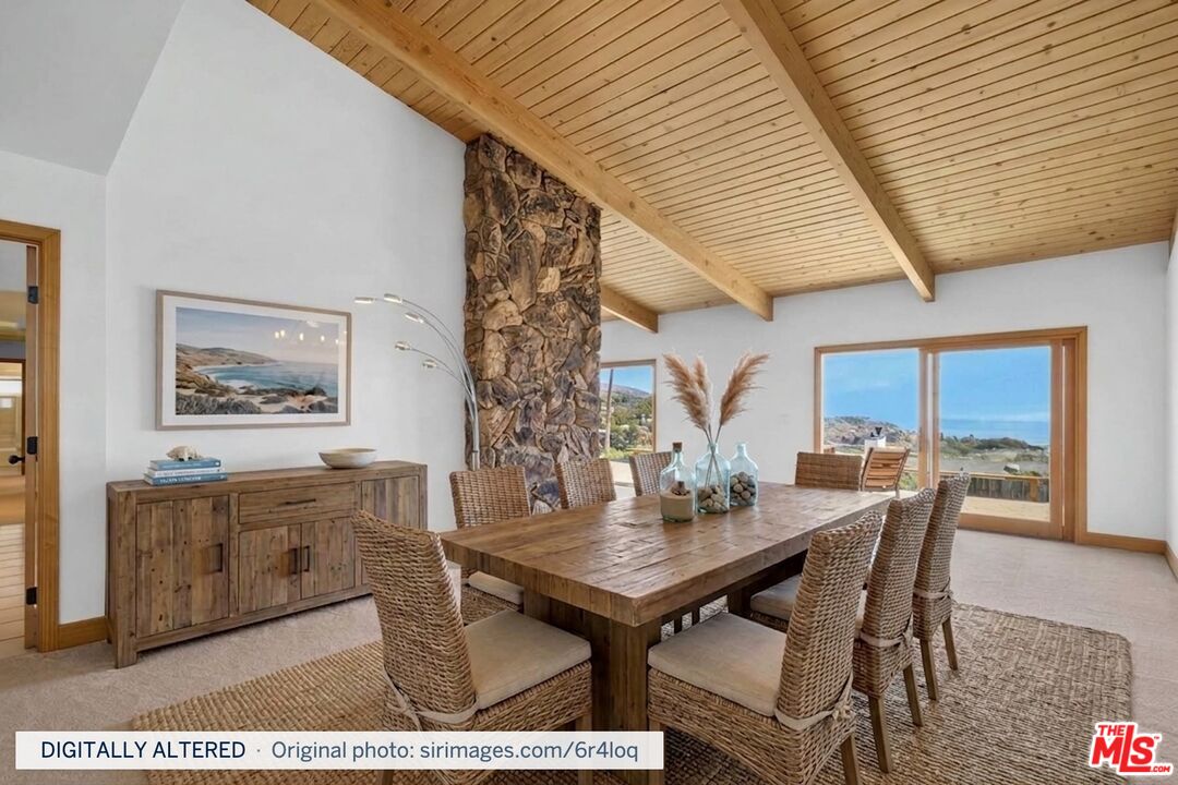 Dining room with a vaulted wood ceiling, stone column, woven chairs, and ocean views through large windows.