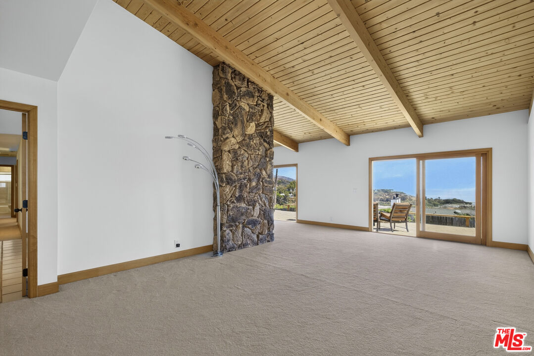 Vacant living room with a vaulted wood ceiling, stone chimney, and sliding doors opening to a coastal view.