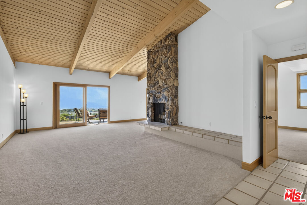 Vacant living room with a vaulted wood ceiling, stone fireplace, and sliding doors to a coastal view.