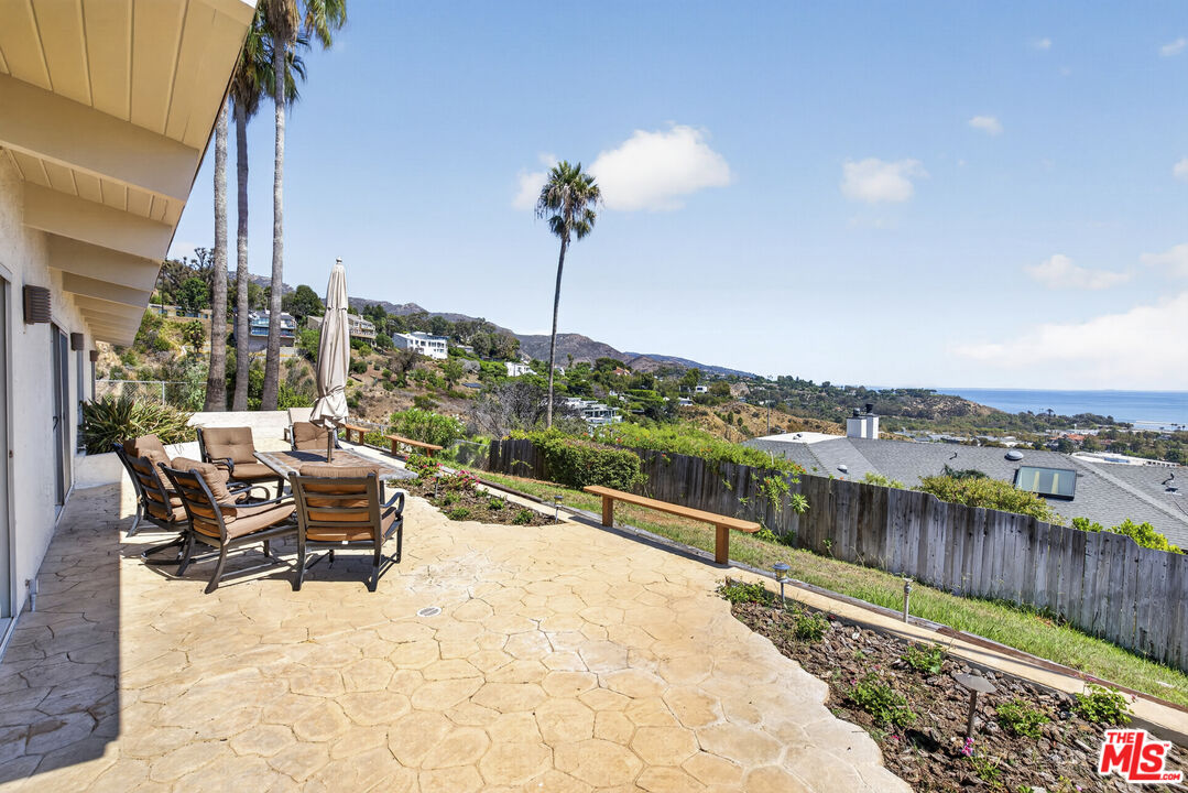 Stone patio with outdoor dining furniture, a tall palm, hillside homes, and a glimpse of the ocean.