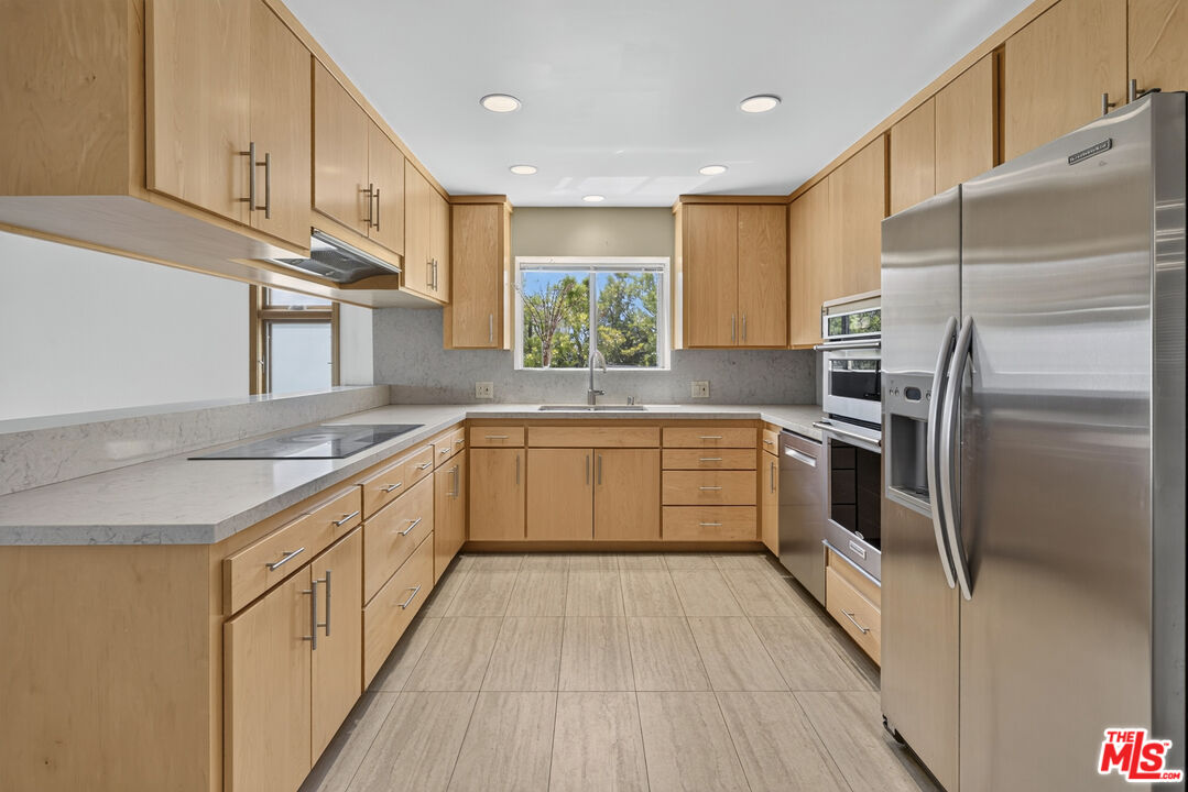 Vacant kitchen with light wood cabinets, pale stone counters, stainless steel appliances, and a window above the sink.