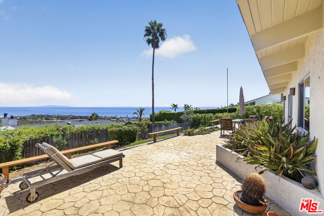 Stone patio with lounge chairs, ocean view, and a tall palm beside the house.