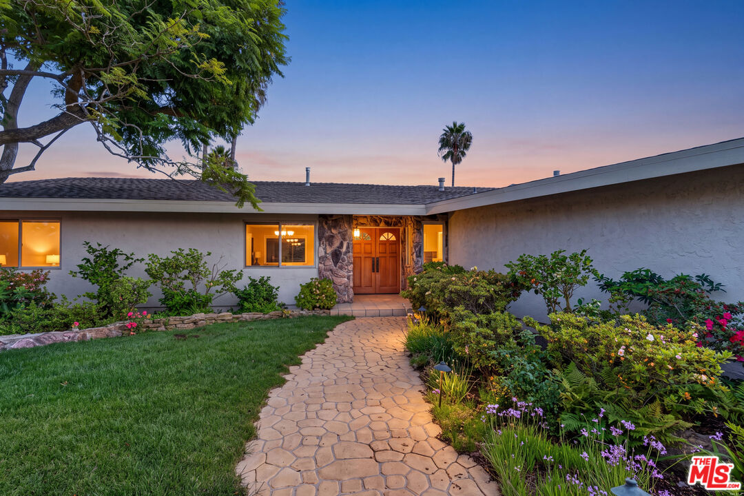 Single-story home with a stone-accented entry and curved walkway amid landscaped gardens at dusk.