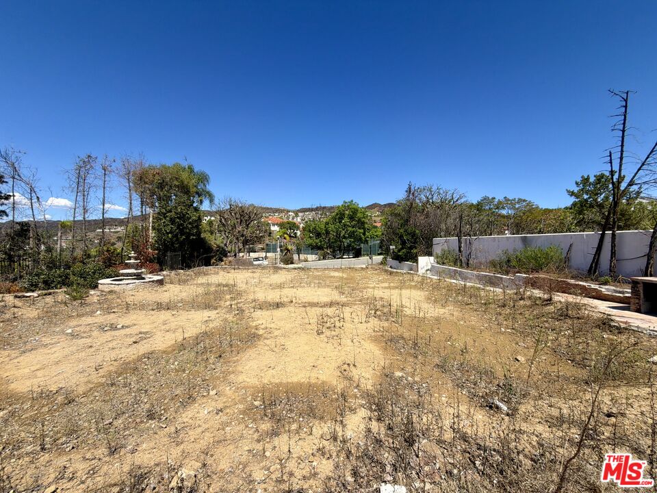 Vacant residential lot with dry bare ground, a white boundary wall, and distant hills.