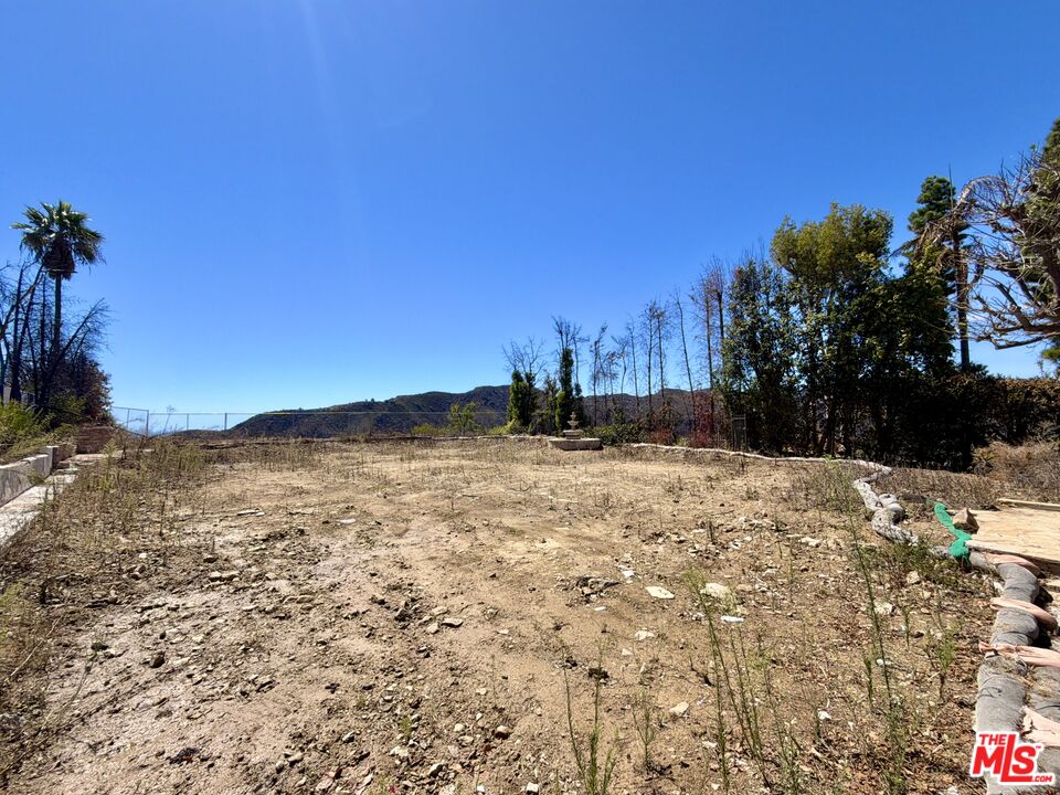 Vacant lot with bare earth, sparse weeds, a chain-link fence, and distant hills.