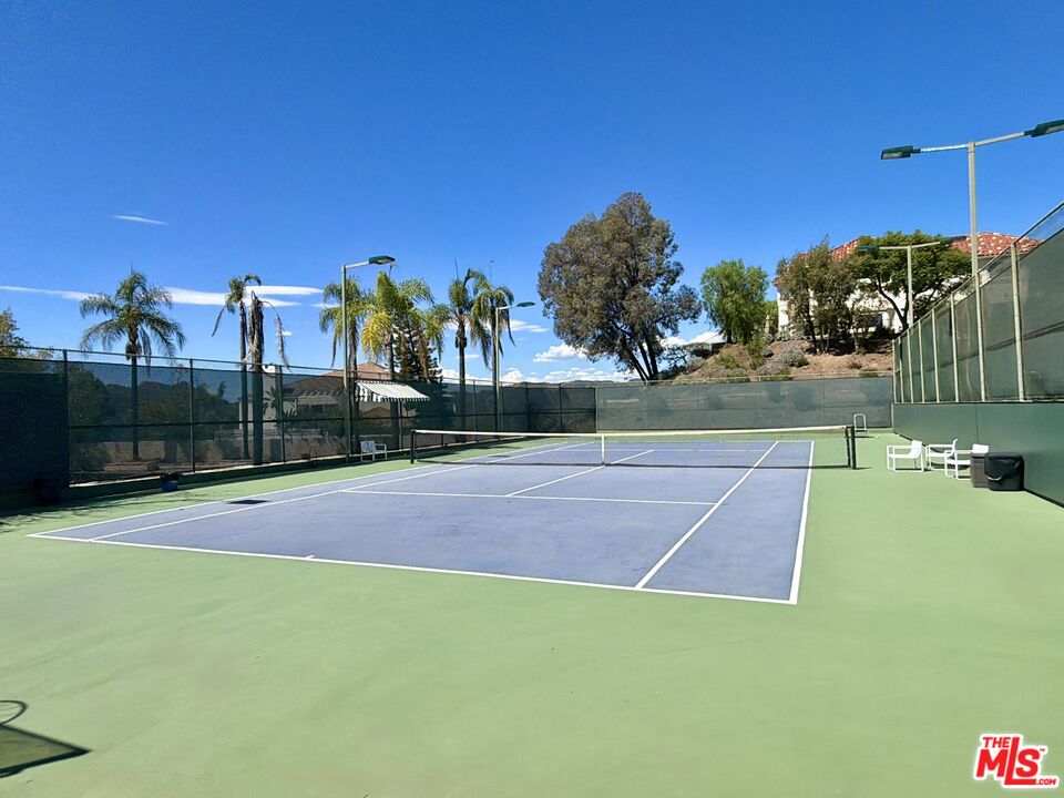Outdoor tennis court with blue playing surface, green perimeter, and surrounding palm trees.