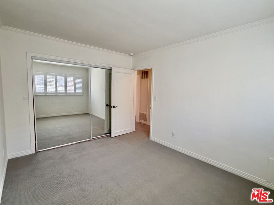 Empty carpeted bedroom with a mirrored sliding closet door and an adjoining room with shuttered windows.
