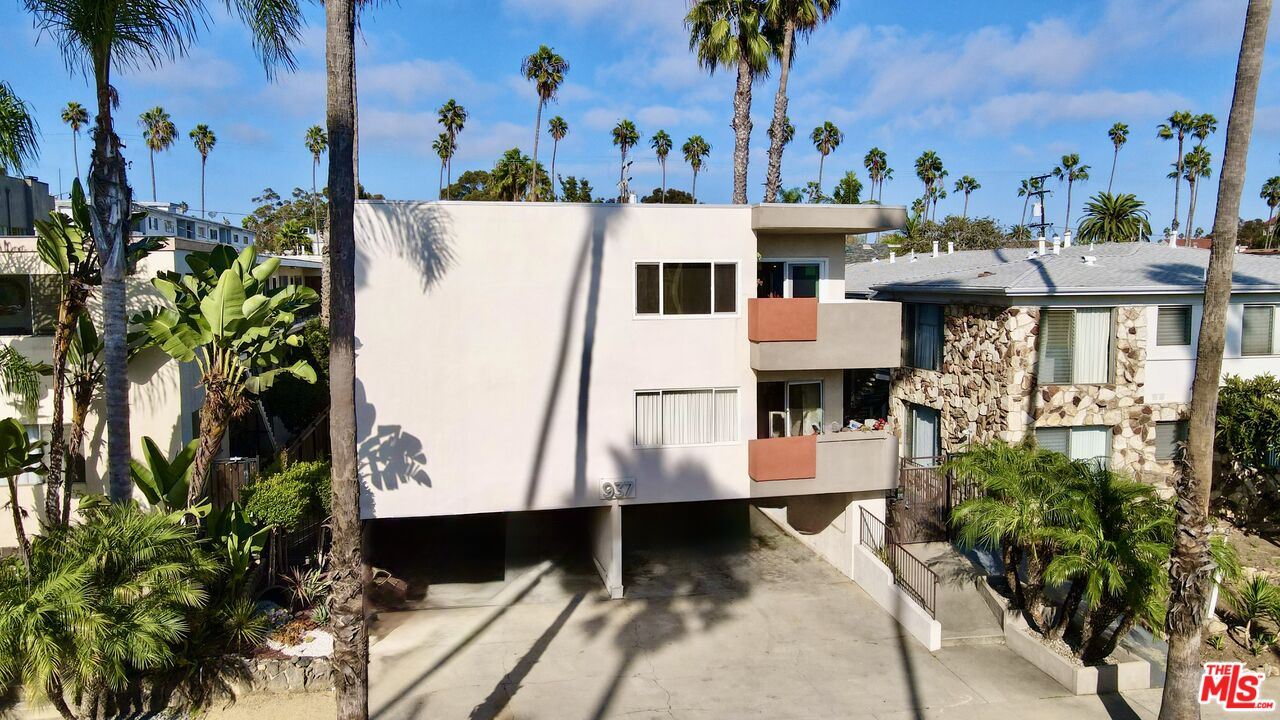 Apartment building with a pale facade and balconies, framed by palm trees above the driveway.