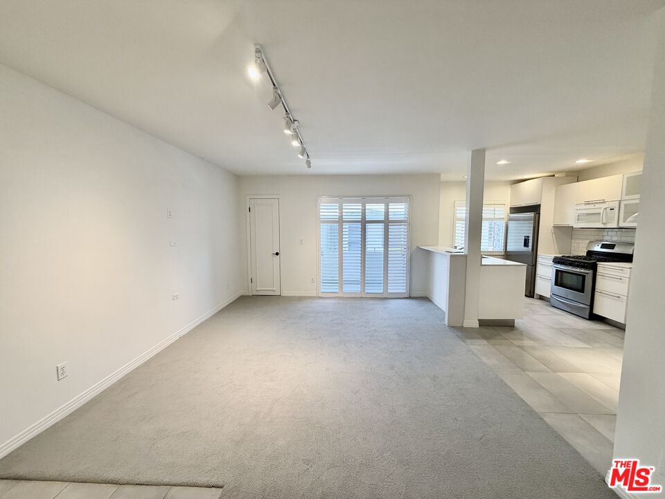 Empty carpeted living room opening to a bright kitchen, with shuttered glass doors and stainless steel appliances.