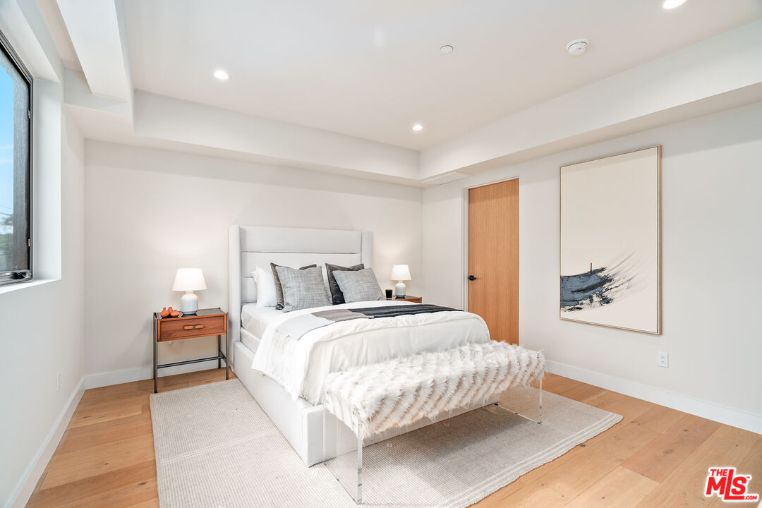 Staged bedroom with a white upholstered bed, fluffy throw, pale wood floors, and abstract wall art.