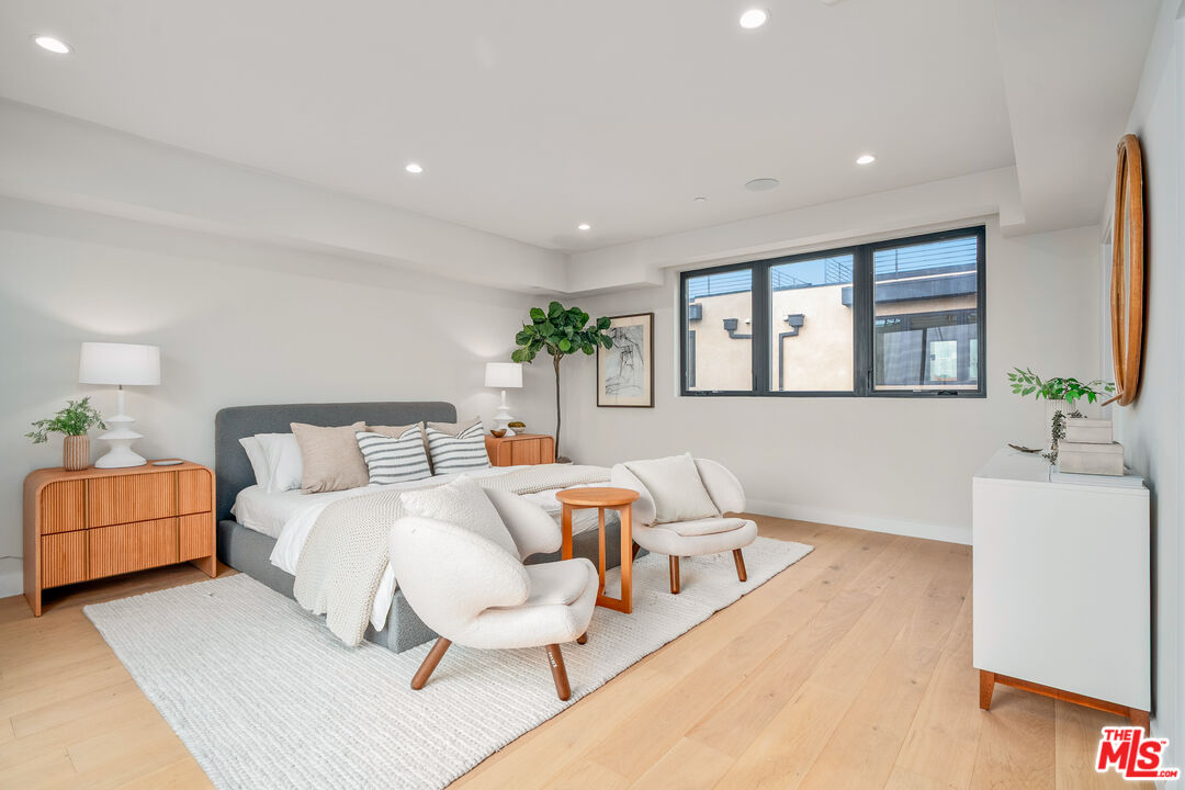 Staged bedroom with a gray bed, cream lounge chairs, and a wide black-framed window.