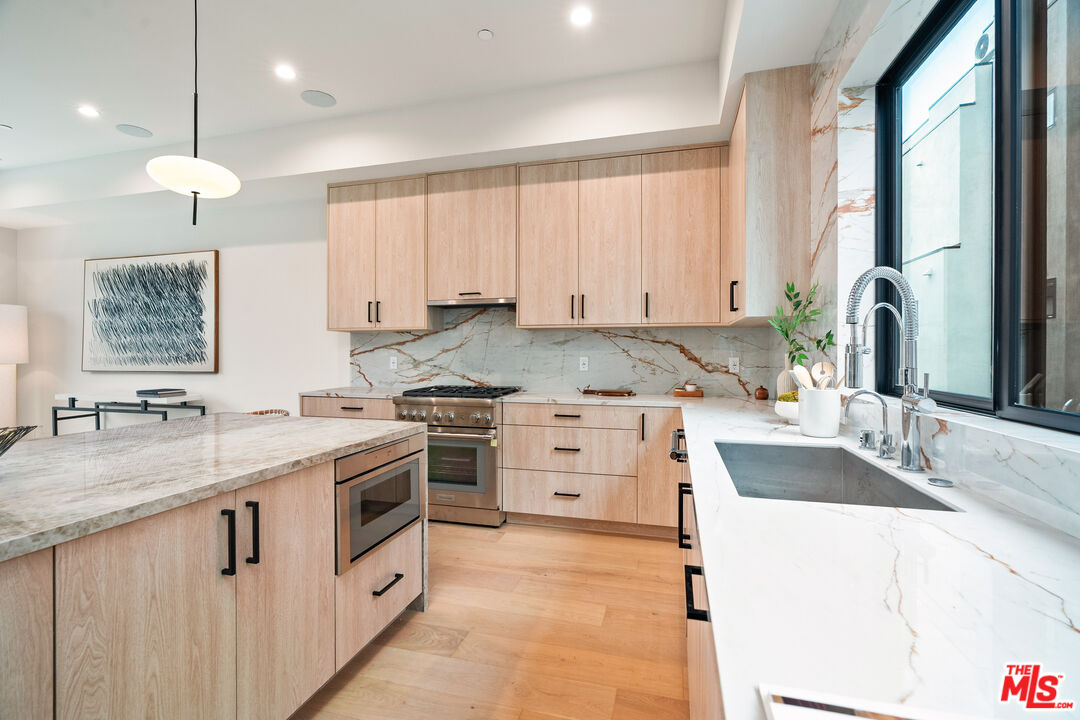 Modern kitchen with light wood cabinets, marble counters, and a large window above the sink.