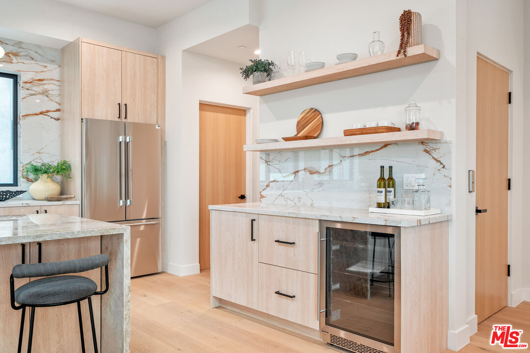 Modern kitchen with light wood cabinets, veined marble counters, open shelves, and a glass-front beverage refrigerator.