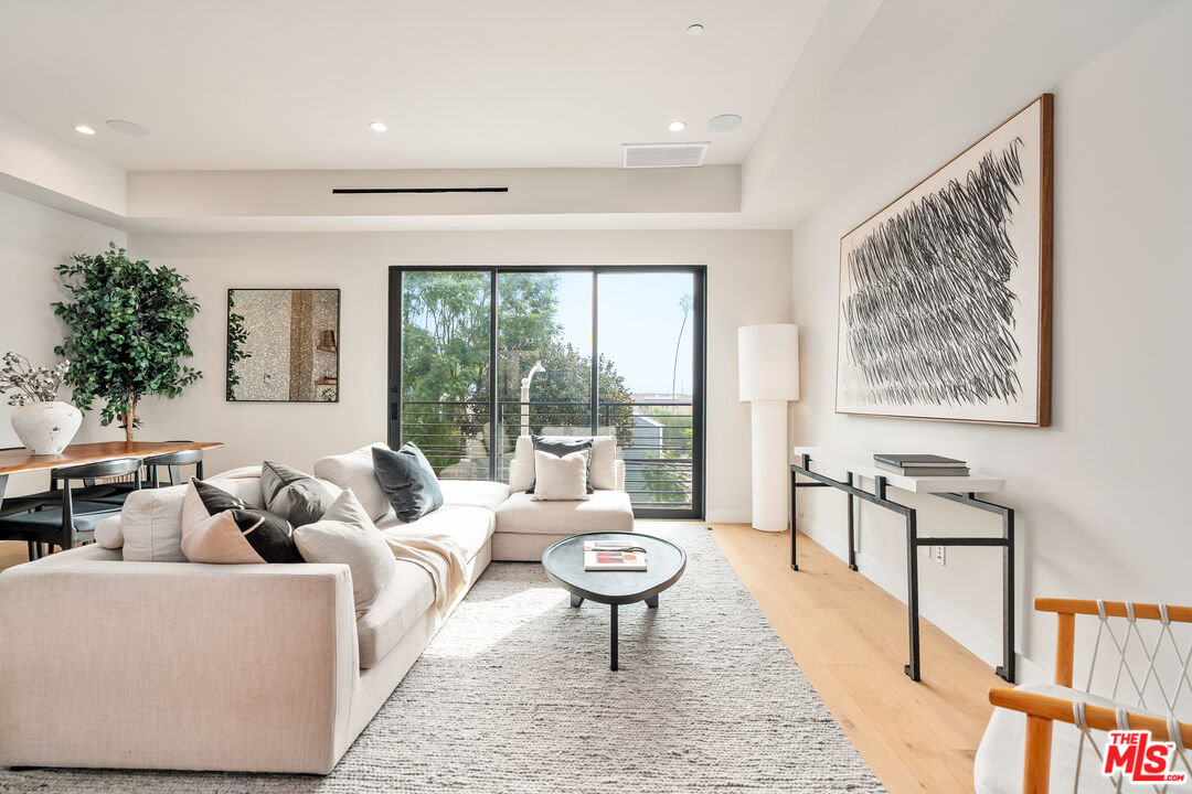 Staged living room with a white sectional, pale wood floors, and sliding doors overlooking leafy views.