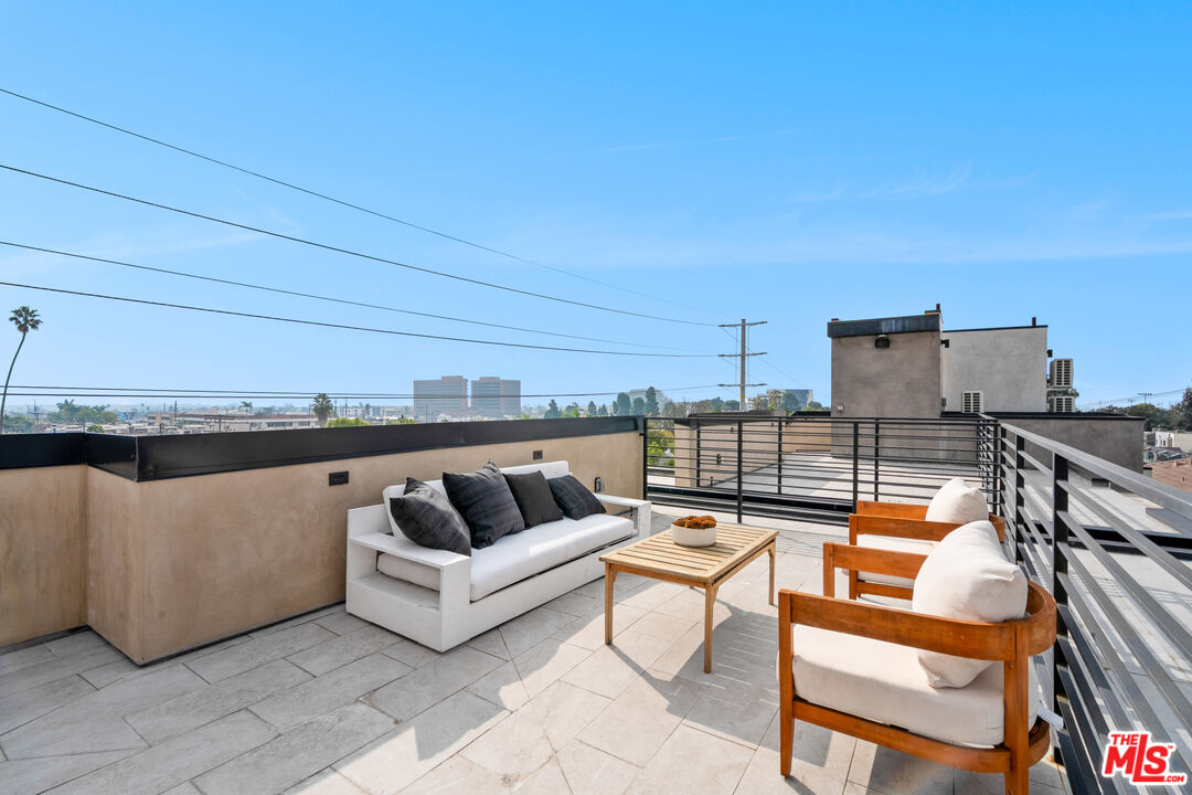 Rooftop terrace with white sofa, cushioned wooden chairs, tiled deck, and distant city view.