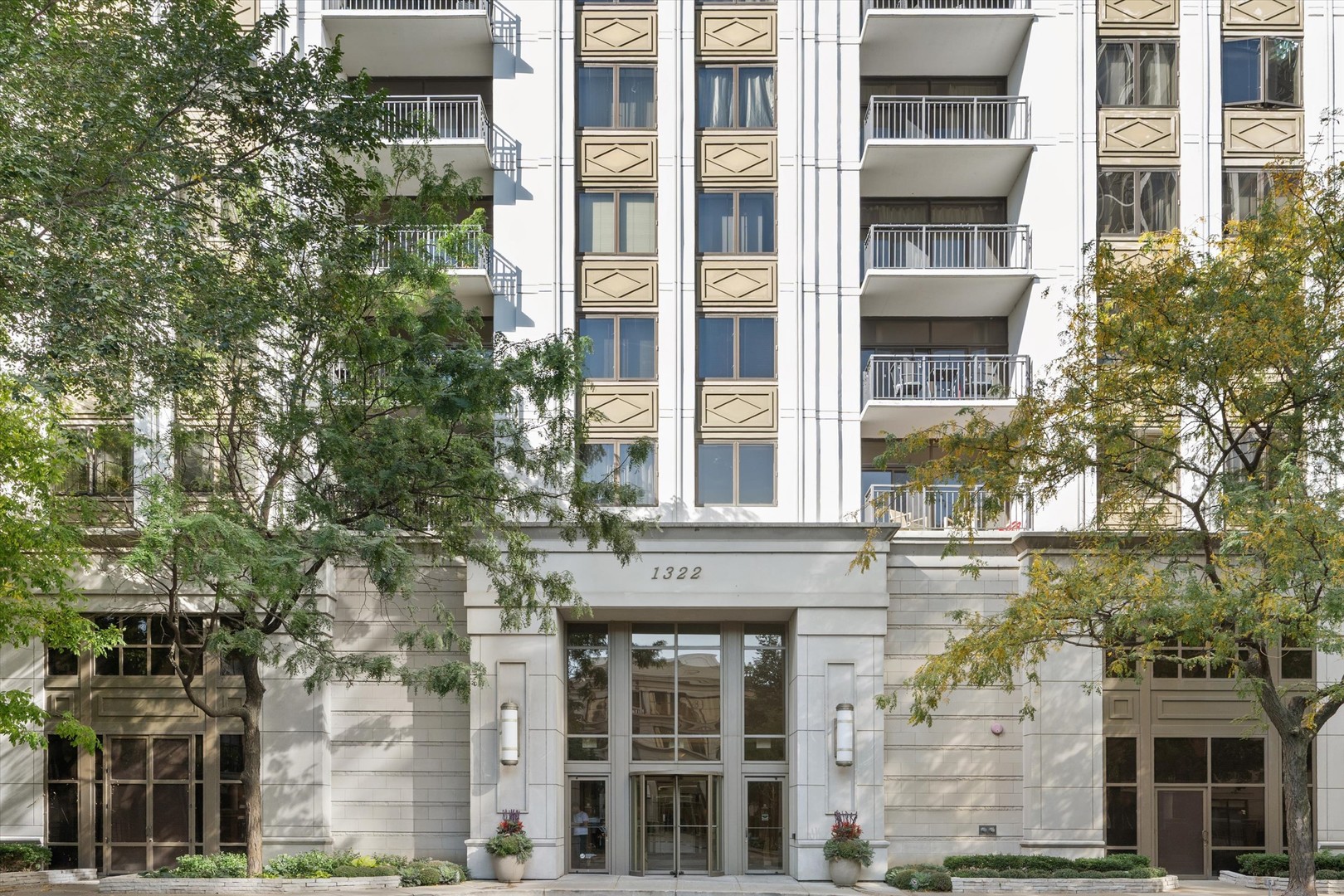 Residential building entrance with glass doors, stone facade, upper balconies, and trees.