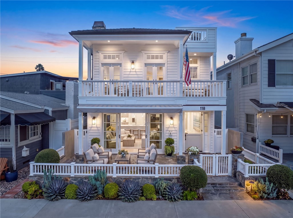 White two-story coastal home with a wide balcony, American flag, and landscaped patio at dusk.