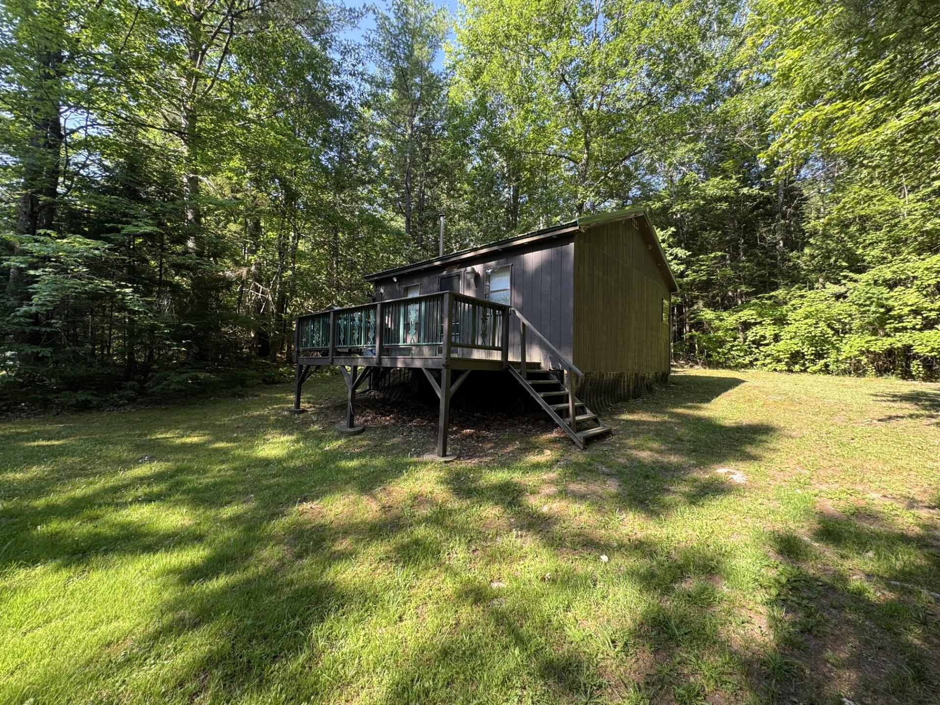 Raised dark-sided cabin with a front deck and stairs in a sunny wooded clearing.