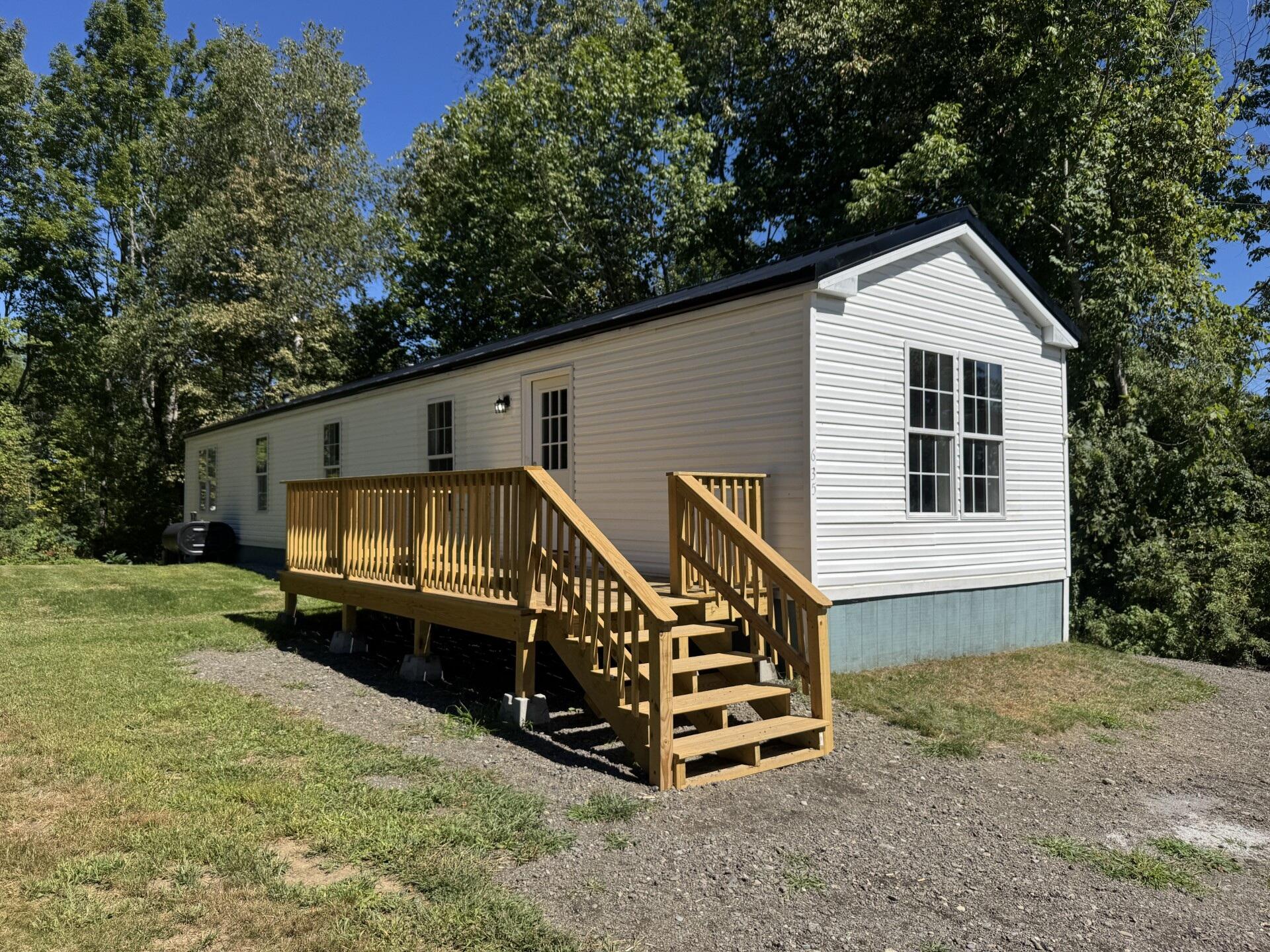 White-sided home with a raised wooden deck and stairs, set among trees beside a gravel drive.