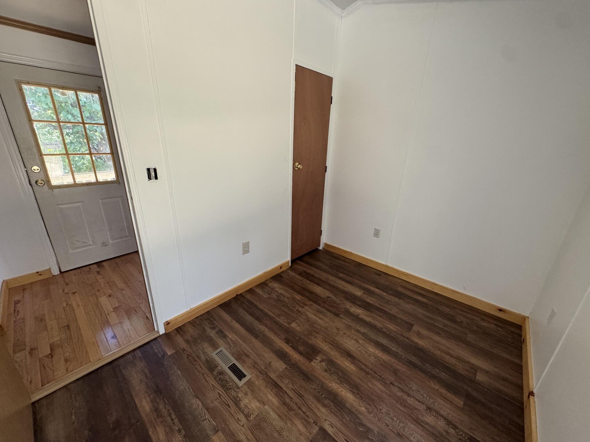 Vacant room with dark plank flooring and wood trim, opening to an entry with a windowed exterior door.