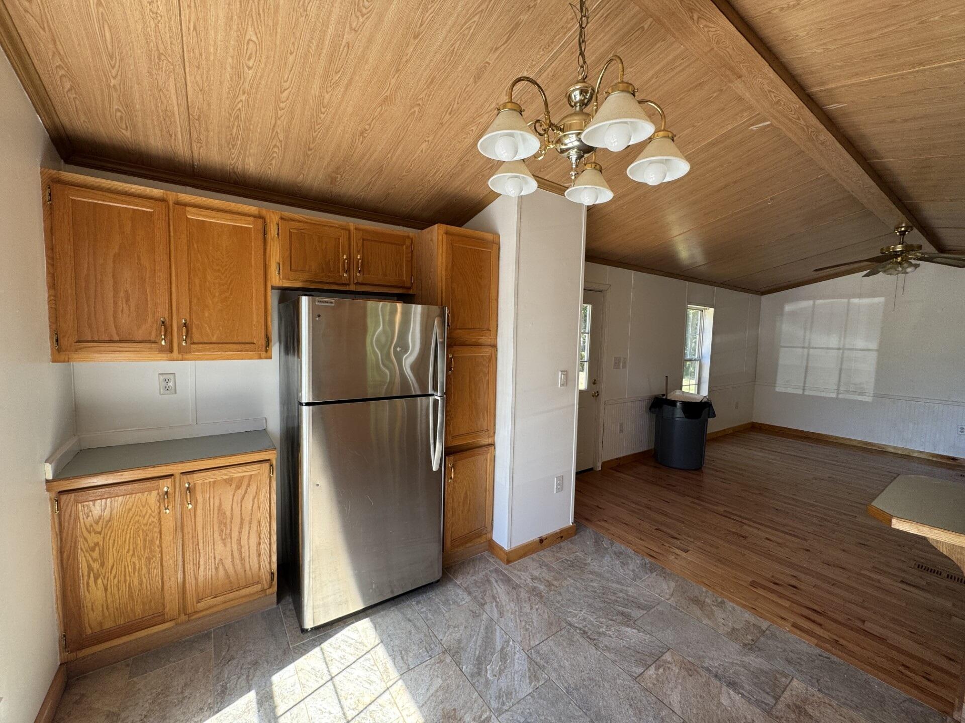 Kitchen with oak cabinets and stainless refrigerator opens into a spacious room beneath a vaulted wood ceiling.