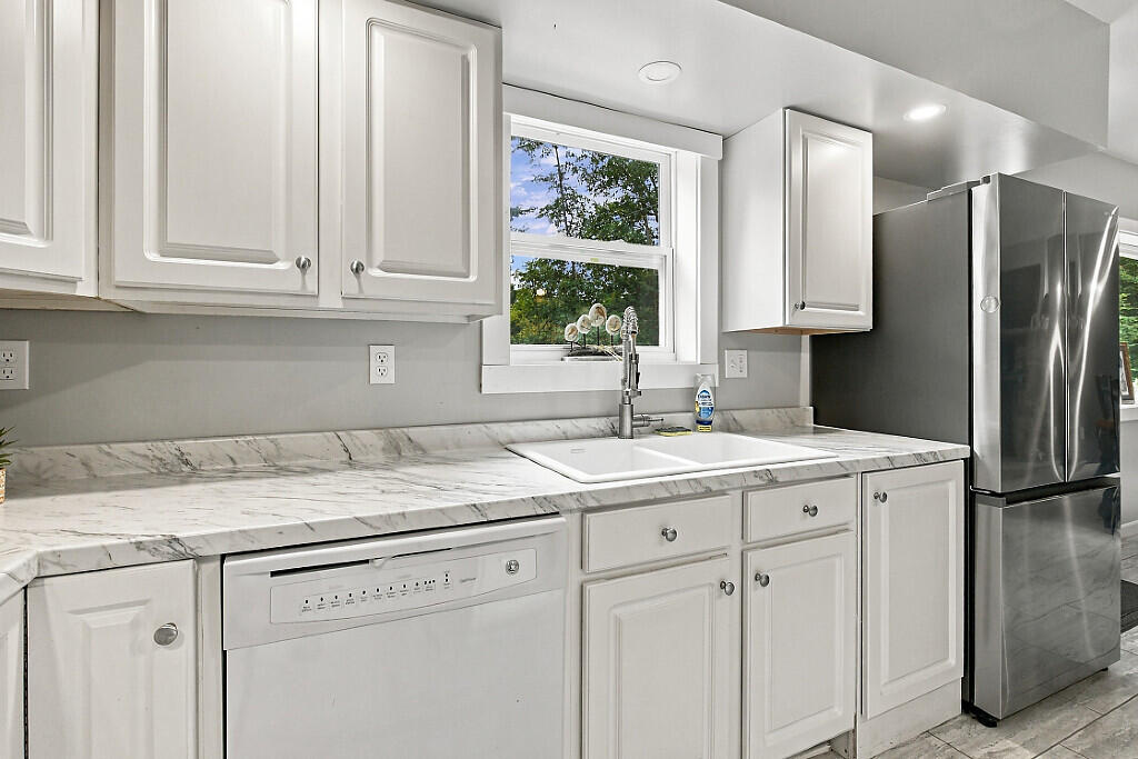 White kitchen with a window above the sink, marble-look counters, dishwasher, and stainless steel refrigerator.