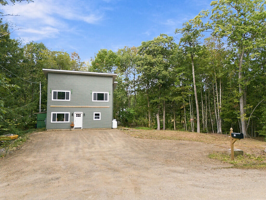 Gray-green two-story house beside a wooded lot, with a broad gravel driveway and roadside mailbox.