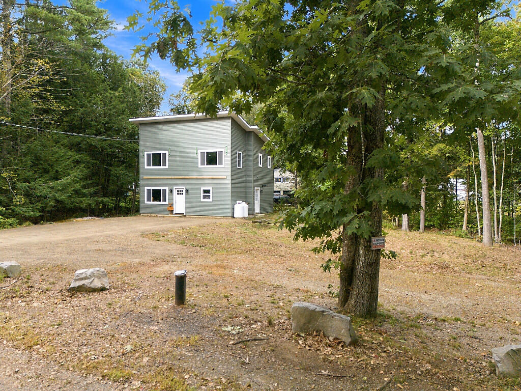 Green two-story house in a wooded gravel clearing, partly obscured by a large tree.