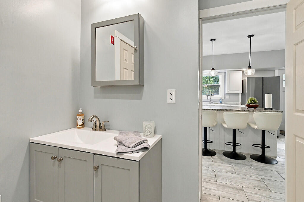 Bathroom vanity with gray cabinetry and folded towels beside an open kitchen doorway.